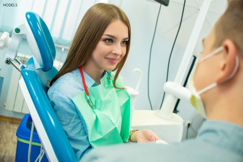 A girl sitting in the dental chair, looking at the dentist talking to her.