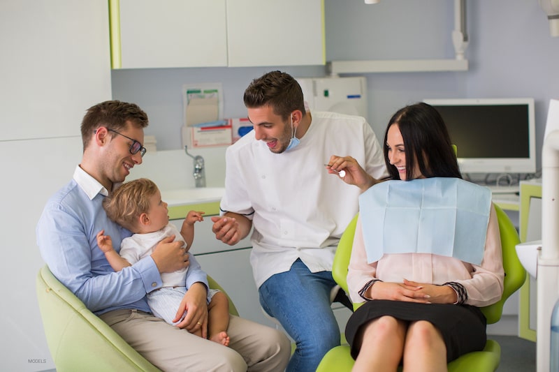 Dad holds baby while the dentist works on mom and shows baby dental tools.