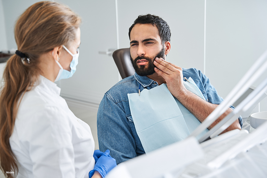 An attractive bearded man in a dentist chair talks to a female dentist while holding his painful jaw.
