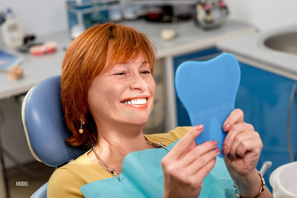 Woman sitting in a dental office holding a mirror and smiling while looking at her teeth.